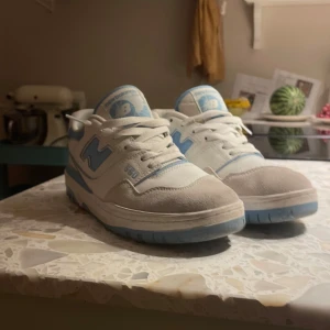 New Balance 550 vit/blå sneakers - Snygga New Balance 550 sneakers i vitt och ljusblått med klassisk siluett. Skorna har ovandel i skinn och mocka. Platt sula och snörning för en clean look. Bra skick, använda några gånger med små smutsiga ytor som försvinner med vatten. Storlek 44!