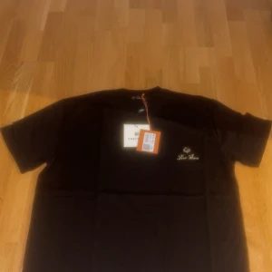 Svart t-shirt från Loro Piana - Svart t-shirt från Loro Piana med broderad logga och emblem på bröstet. Klassisk passform med rund hals och korta ärmar. Materialet känns mjukt och lyxigt, perfekt för dig som gillar stilrena och exklusiva plagg.