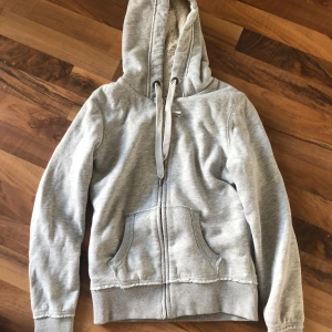 Grå hoodie med teddyfoder från H&M - Mysig grå hoodie från H&M L.O.G.G. med fluffigt teddyfoder i hela insidan. Tröjan har dragkedja, huva med snörning och två stora fickor framtill. Perfekt för kyliga dagar när du vill vara både varm och chill.