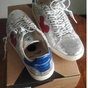 Disney x MOA glittriga sneakers - Snygga sneakers med silverglittriga paljetter, röd Mickey Mouse-detalj på sidan och blå metallic häl. Skorna har vita snören med svart text och en klassisk rund tå. Perfekta för dig som vill sticka ut med en lekfull och trendig stil.