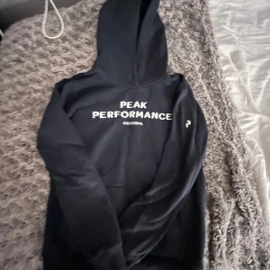 Svart hoodie från Peak Performance - Svart hoodie från Peak Performance med vit logga och text på bröstet. Klassisk modell med huva och känguruficka, perfekt för chill dagar. Mjuk insida och ribbade muddar. Snygg och enkel stil som funkar året runt.💕