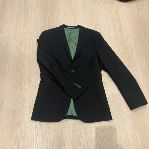 Svart kavaj J.Lindeberg slim fit S - Stilren svart kavaj från J.Lindeberg i slim fit-modell. Kavajen har klassisk passform med två knappar framtill, fickor med lock och grön insida i glansigt material 3 fickor. Perfekt för dig som vill ha en snygg och modern look. 