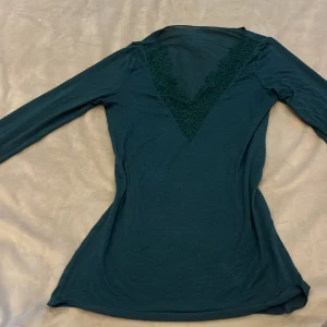 Mörkgrön topp med spets från Intimissimi - Supersnygg mörkgrön långärmad topp från Intimissimi med v-ringning och spetsdetaljer både vid halsen och ärmsluten. Tunn och mjuk i materialet, 😊