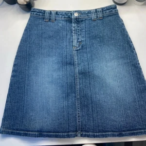 Blå jeanskjol med knappar från Helline - Snygg blå jeanskjol från Helline med klassisk A-linjeform, bälteshällor och två bakfickor med lock och knapp. Kjolen är i stretchig denim med 96% bomull och 4% elastan, perfekt för en avslappnad och cool look.