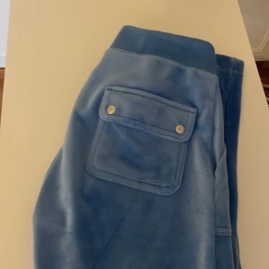 Blå mjukisbyxor Juicy Couture XXS - Blåa mjukisbyxor från Juicy Couture i supermjuk velour med broderad JC-logga och ficka med tryckknappar bak. Klassisk Juicy-detalj i metall på snöret. Perfekta för en chill och trendig look. Jag har även storlek XS i vanliga kläder men i mina juicy har jag XXS då dom är jätte långa. Jag kan gå med på att sänka pris. 