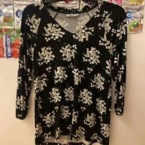 Svart blommig topp från me&i - Svart långärmad topp från me&i med vitt blommönster och gröna detaljer. V-ringad med volangkant vid halsen och elastiska muddar vid ärmslut. Skön passform och perfekt för dig som gillar en söt och chill stil.