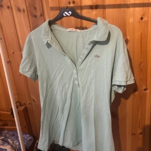 Ljusgrön pikétröja från Lacoste - Snygg ljusgrön/turkås pikétröja från Lacoste med klassisk krage och knappar framtill. Tröjan har korta ärmar och den ikoniska krokodilloggan broderad på bröstet. Perfekt för en fräsch och sportig look under varmare dagar.