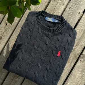 Mörkblå kabelstickad tröja Polo Ralph Lauren - Kabelstickad mörkblå tröja från Polo Ralph Lauren med klassisk rund hals och röd broderad logga på bröstet. Tröjan är i bomull och har ribbade muddar vid ärmslut och nederkant. Perfekt för dig som gillar stilren och tidlös design. Storlek L men sitter som M