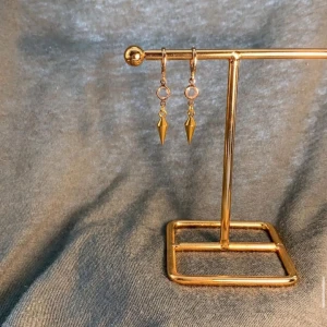 Guldiga örhängen med spetsig hängande detalj - Snygga örhängen i guld med rund ögla och en liten transparent sten. Nedanför hänger en spetsig, konformad detalj som ger ett edgy intryck. Perfekta för dig som vill sticka ut med en cool accessoar.