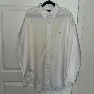 Vit skjorta från Ralph Lauren - Klassisk vit skjorta från Ralph Lauren i bomull med button-down krage och broderad logga på bröstet. Skjortan har lång ärm, knappar framtill och en tidlös, clean look. Classic fit-modell som passar till många olika stilar. As najs under sommaren! 