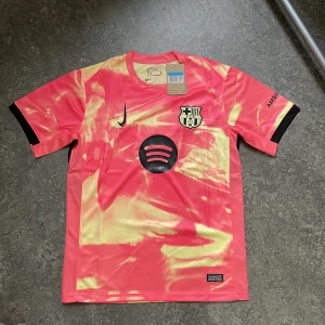 FC Barcelona Tredjetröja 2023/24 – Rosa/Beige (Storlek M) - Limited edition FC Barcelona tredjetröja säsongen 2023/24 – med unikt färgblocksmönster i rosa och beige samt svart Nike-logga och Spotify-sponsor. Ett färgstarkt och ovanligt plagg som sticker ut både på läktaren och i vardagen!  ✔️ Säsong: 2023/24 ✔️ Storlek M – Slim fit ✔️ I nyskick – aldrig använd