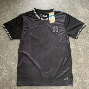 Brasilien Shadow Camo (Nike) – Rare - Unik Brasilien-tröja i helsvart camo-look. Ton-i-ton-logga och diskreta detaljer ger en riktigt clean och edgy look. Passar både till vardags och sport. 🧼 Nytt skick 📏 Storlek: 1M 💎 Svår att hitta – perfekt för dig som vill sticka ut