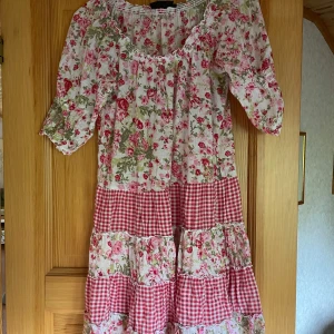 Blommig sommarklänning  - Supersöt långklänning från KappAhl med blommigt och rödvitt rutigt mönster. Klänningen har trekvartsärm, volangdetaljer och en romantisk offshoulder-ringning. Perfekt för sommarens alla tillfällen. Lätt och luftig i bomull.