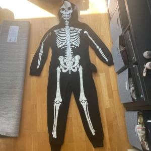Svart hoodie med skelettmönster och huva - Passar män som är över 180 cm!! Svart hoodie i onesie-stil med vit skelettprint över hela kroppen och huva med döskalletryck. Långärmad och dragkedja framtill. Perfekt för halloween eller temafest, riktigt skön och unik look.
