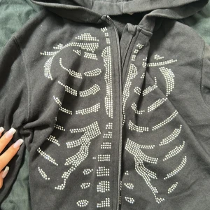Svart hoodie med skelettmönster - Svart hoodie med huva och dragkedja framtill. Framsidan är dekorerad med ett skelettmönster i silvriga nitar som ger en edgy vibe. Perfekt för dig som gillar unika detaljer och streetstyle. Mjuk och skön, passar till många outfits. Storlek 146-156