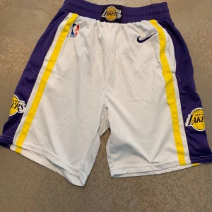 Lakers basketshorts från Nike - Snygga basketshorts från Nike med Los Angeles Lakers-logga. Shortsen är vita med lila och gula sidopaneler, elastisk midja och broderade detaljer. Perfekta för basket eller streetwear. Klassisk NBA-stil med coola färgblock och officiella märken.