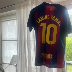 FC Barcelona Lamine Yamal tröja Nike M - Säljer en officiell FC Barcelona fotbollströja från Nike i storlek M. Tröjan har Lamine Yamal och nummer 10 på ryggen, klassiska blå och röda vertikala ränder, gult Nike-märke och Spotify-logga på bröstet. Tillverkad i lätt, ventilerande material perfekt för match eller träning.