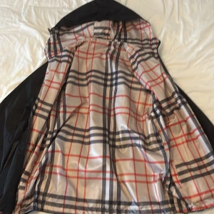 Svart vindjacka Burberry med rutig lining - Svart vindjacka från Burberry med klassiskt rutig insida i beige, rött och svart. Jackan har huva, dragkedja framtill och logga på bröstet. Perfekt för dig som vill ha en stilren jacka med lyxig känsla och snygga detaljer.