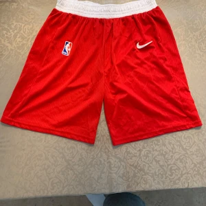 Röda NBA Nike shorts med vit resår Chicago Bulls - Snygga röda basketshorts från Nike med NBA-logga på ena benet och vit elastisk midja. Klassisk loose fit och sportig look, perfekta för träning eller basket. Materialet är lätt och andas, med diskreta ränder i tyget och broderad Nike Swoosh.