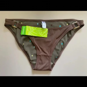 Bruna bikiniunderdelar  - Snygga bikiniunderdelar i brun färg från Mix & Match. Insidan har ett lekfullt prickigt mönster i blått, gult och brunt. Sidorna har justerbara spännen i plast för perfekt passform. Materialet är mjukt och stretchigt, perfekt för strandhäng.