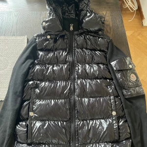 Svart pufferjacka från Moncler - Säljer en svart pufferjacka från Moncler med glansig finish och stickade svarta ärmar. Jackan har huva, dragkedja framtill, fickor med tryckknappar och en ikonisk Moncler-logga på ärmen. Perfekt för kyliga dagar när du vill ha både stil och värme.