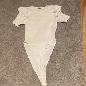 Vit ribbad bodysuit med volangärm Zara - Supersnygg vit bodysuit från Zara med ribbad struktur och korta ärmar med volangdetaljer på axlarna. Rund halsringning och figurnära passform. Perfekt att styla med jeans eller kjol för en trendig look.
