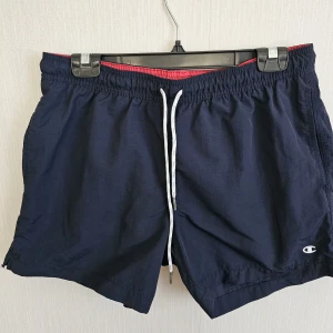 Mörkblå shorts från Champion - Mörkblå shorts från Champion med vit snörning i midjan och röd insida. Klassisk loose passform, sidofickor och bakficka. Perfekta för sommaren och träning, tillverkade i lätt polyester för en skön känsla.