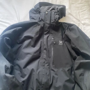Svart vindjacka från Haglöfs XL - Svart vindjacka från Haglöfs i storlek XL. Jackan har huva, dragkedja framtill och en bröstficka med dragkedja. Tillverkad i återvunnen polyester med vattentät PROOF-membran. Perfekt för blåsiga och regniga dagar.