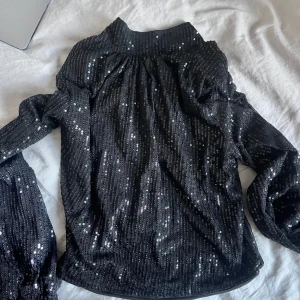 Svart paljettopp med öppen rygg - Glittrig svart topp med långa ärmar och öppen rygg. Toppens yta är täckt av små paljetter som ger ett riktigt snyggt party-vibe. Perfekt för dig som vill sticka ut och glänsa. Sitter snyggt med lite loose fit och har hög hals framtill. Använd en gång 