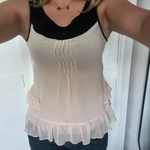Beige volangtop med svarta detaljer - Säljer en söt beige linnetopp med svarta axelband och kant upptill. Toppen har volangdetaljer längs nederkanten och sidorna samt veck framtill för extra stil. Jag har själv figursytt denna med symaskin. Om ni har frågor är det bara att skriva💕💕