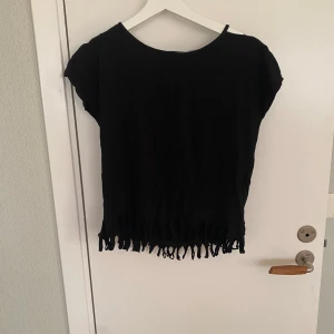 Svart topp med fransar och öppen rygg - Cool svart topp med korta ärmar, öppen rygg och fransar längs nederkanten. T-shirten har en avslappnad passform och är tillverkad i mjukt bomullstyg. Perfekt för dig som vill ha en edgy och unik stil.