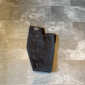 Svarta jeans från Nudie Jeans - Svarta jeans från Nudie Jeans i modellen Tight Long John. Klassisk femficksdesign, smal passform och stretchigt bomullstyg. Perfekta svarta jeans med snygg siluett och diskreta detaljer. Storlek 32/34.