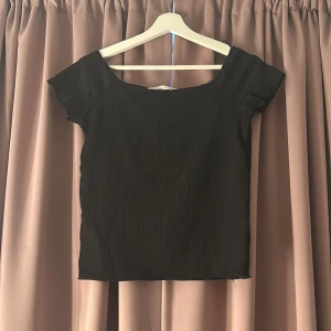 Svart ribbad topp med kort ärm - En svart topp med ribbad struktur och korta ärmar. Toppen har en bred, något båtformad halsringning och är figurnära utan att vara tight. Perfekt att styla med jeans eller kjol för en clean look.🖤