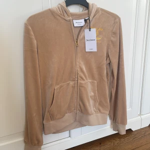 Beige velourjacka Juicy Couture - Supermjuk beige velourjacka från Juicy Couture med huva och dragkedja. Guldig logga på bröstet och stor Juicy Couture-text i gult på ryggen. Jackan har kängurufickor och ribbade muddar för en chill och trendig look. Är helt oanvänd.