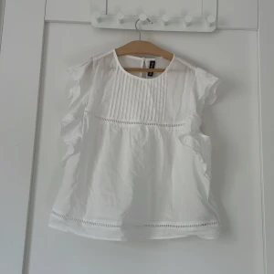 Vit blus med volang från H&M - Superfin vit blus från H&M med volangdetaljer längs ärmarna och smala veck framtill. Blusen är ärmlös med rund halsringning och har dekorativa broderade band både upptill och nertill. Perfekt för en fräsch och luftig look.