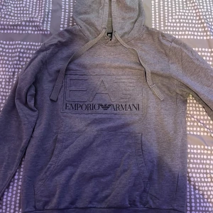 Grå hoodie från Emporio Armani EA7 - Snygg grå hoodie från Emporio Armani EA7 med stor logga framtill. Tröjan har huva med snörning och en klassisk känguruficka. Perfekt för dig som gillar stilrena och exklusiva streetwear-plagg.