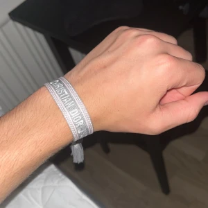 Grått Christian Dior armband i tyg - Snyggt armband från Christian Dior i ljusgrått tyg med vävd logga och tofsar i ändarna. Justerbar knytning gör att det passar de flesta handleder. Perfekt accessoar för att lyfta din outfit med en exklusiv touch.