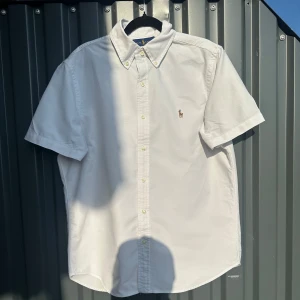 Vit kortärmad skjorta Ralph Lauren M - Vit kortärmad skjorta från Ralph Lauren i klassisk passform. Skjortan har button-down krage, broderad logga på bröstet och knäppning framtill. Tillverkad i bomull och perfekt för en clean och stilren look. Passar en som är mellan 170-180