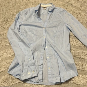 Ljusblå skjorta med knappar - Klassisk ljusblå skjorta med lång ärm och knäppning framtill. Skjortan har en enkel och stilren design med krage och vita knappar. Perfekt för en clean och fräsch look. Tillverkad i mjukt bomullstyg som känns skönt mot huden.