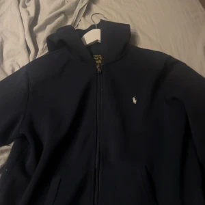 Mörkblå hoodiejacka från Polo Ralph Lauren - Mörkblå hoodiejacka från Polo Ralph Lauren med dragkedja framtill och klassisk vit logga på bröstet. Jackan har huva och två fickor, perfekt för en chill och stilren look. Tillverkad i mjuk bomull för extra komfort.