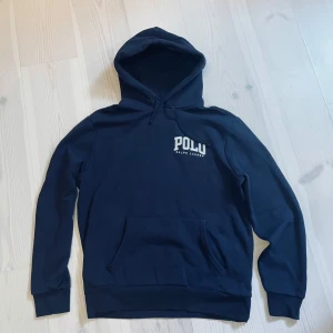 Hoodie | RALPH LAUREN. - Snygg marinblå hoodie från Polo Ralph Lauren med vit logga på bröstet. Klassisk känguruficka framtill, huva med snörning och ribbade muddar. Perfekt för en chill och stilren look. (OANVÄND) (ORIGINALPRIS: 1607kr).