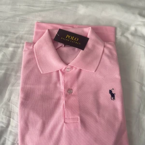 Rosa pikétröja från Polo Ralph Lauren - Snygg rosa pikétröja från Polo Ralph Lauren med klassisk krage och två knappar framtill. Tröjan har korta ärmar och den ikoniska broderade loggan i mörkblått på bröstet. Perfekt för en fräsch och sportig look.