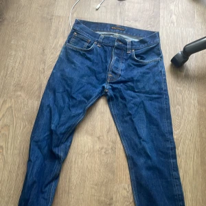 Blå jeans från Nudie Jeans - Säljer ett par klassiska blå jeans från Nudie Jeans med snygga kontrastsömmar och ikoniska bakfickor med vågig brodyr. Jeansen är i 100% bomull och har en rak passform med normal midja. Perfekta för dig som gillar stilrena och tidlösa jeans.