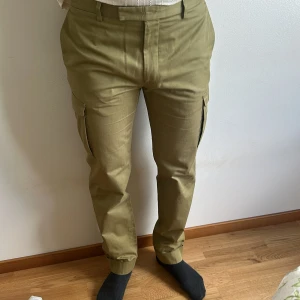 Olivgröna Gant-byxor med raka ben - Säljer ett par olivgröna cargopants från Gant med raka ben och klassiska cargofickor på sidorna. Byxorna har bälteshällor, knäppning fram och bakfickor med knapp. passformen är normal med lite tapered ben. 100% bomull