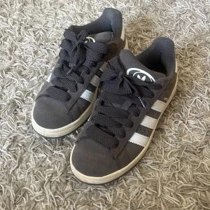 Adidas Campus sneakers i grå mocka - Snygga Adidas Campus sneakers i mörkgrå/ beige mocka med klassiska vita ränder på sidorna och vit logga på hälen. Skorna har rund tå, platt sula och breda svarta snören. Perfekta för en streetstyle-look och funkar året runt. Köpa från JD men finns inte så mycket täcken på användning. 