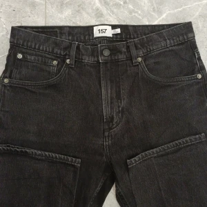 Svarta cargopants från 157, W34 L32 - Svarta cargopants från 157 med klassisk femficksdesign och stora fickor på benen. Jeansen har raka ben och är tillverkade i robust denim. Perfekta för dig som gillar en avslappnad och stilren look.