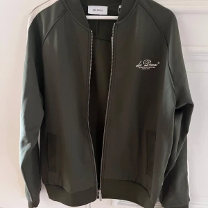 Olivgrön track jacket från Les Deux - Snygg olivgrön track jacket från Les Deux med vit logga på bröstet och dragkedja framtill. Jackan har ribbade muddar och en clean design med vita detaljer på axlarna. Perfekt för en sportig och stilren look.