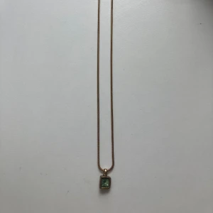 Guldhalsband Pilgrim med grön sten - Snyggt guldhalsband från Pilgrim med en fyrkantig grön sten i ett stilrent hänge. Kedjan är tunn och elegant, perfekt för att ge en touch av färg till din look. Märkt med Pilgrim-tag vid låset.