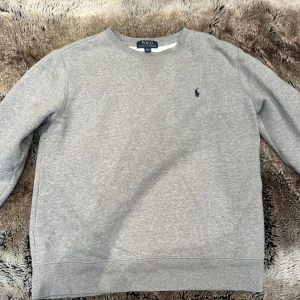 Grå sweatshirt från Polo Ralph Lauren - Klassisk grå sweatshirt från Polo Ralph Lauren med rund hals och broderad marinblå logga på bröstet. Tröjan har långa ärmar, ribbade muddar och är tillverkad i mjuk bomullsmix som är perfekt för chill dagar. Köpt på kidsbrandstore