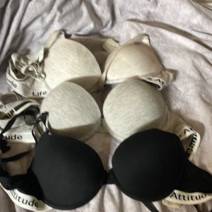 1 vit/grå, 1 grå och 1 svart BH (70B) - Tre bandeau toppar med vadderade kupor, två i ljusgrått och en i svart. Alla har breda axelband med texten 'Life Attitude' i svart. Snygg och sportig look, perfekt att bära under skjorta eller hoodie.